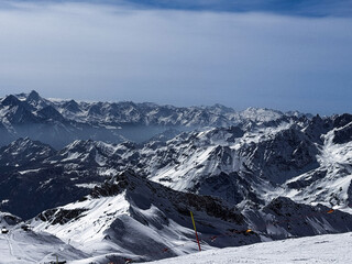 Winter panoramic view of the snowy Alpine mountain range with dramatic peaks and deep valleys under a clear blue sky. Scenic high-altitude landscape ideal for travel and nature themes.