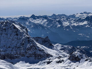 Winter panoramic view of the snowy Alpine mountain range with dramatic peaks and deep valleys under a clear blue sky. Scenic high-altitude landscape ideal for travel and nature themes.