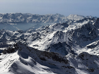 Winter panoramic view of the snowy Alpine mountain range with dramatic peaks and deep valleys under a clear blue sky. Scenic high-altitude landscape ideal for travel and nature themes.