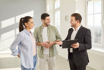 Friendly real estate agent presenting new property to happy couple in modern apartment. Smiling man and woman listening attentively while discussing purchase details with realtor in spacious room.