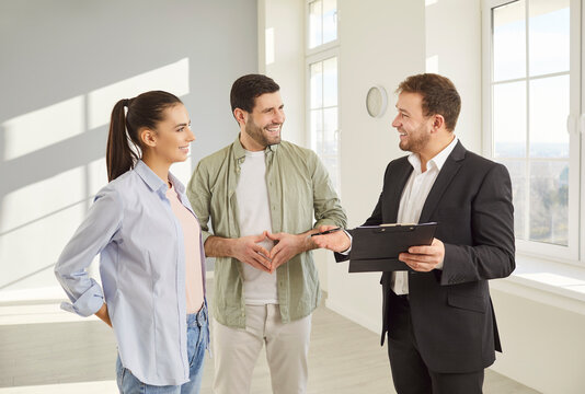 Friendly real estate agent presenting new property to happy couple in modern apartment. Smiling man and woman listening attentively while discussing purchase details with realtor in spacious room.