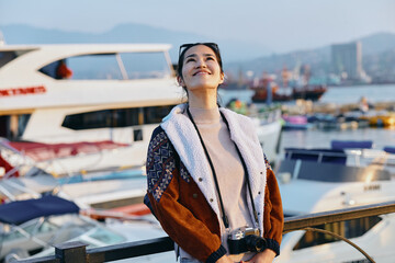 Stylish woman in a warm coat stands beside a marina with boats, sunny harbor backdrop, lifestyle photography with fashion flair and travel vibe, casual elegance by water