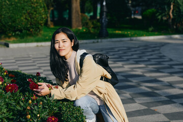 Woman kneeling beside blooming flowers in a park, enjoying a sunny day with a backpack, casual coat, and relaxed pose, capturing natural light, greenery, and outdoor lifestyle in a candid scene