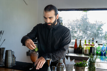 Bartender placing mint garnish on cocktail in hotel bar with greenery.