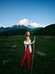 Naklejka premium Woman standing in a green meadow with a wooden staff, red pants and white top, mountains and blue sky behind for an outdoor nature scene