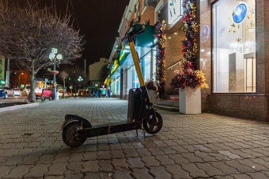 An electric scooter is parked on a paved city sidewalk at night. Shop windows and streetlights illuminate the modern urban scene beautifully. - Powered by Adobe