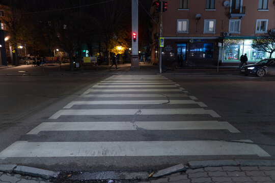 An empty pedestrian crossing in a bustling city street at night with a red traffic light, illuminated by streetlights and shop windows, waiting for pedestrians.