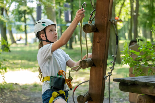 Young girl child in a white safety helmet climbing in a rope adventure park, attaching a carabiner hook to the cable. Kids summer outdoor recreation, obstacle course activity simple concept, sunny day
