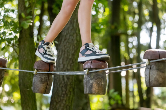 Child's legs in sneakers balancing on unstable wooden mushroom shaped steps, closeup, detail. Rope adventure park obstacle course, walking on a cable, outdoor activity, agility skills sport concept
