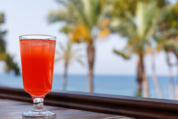 Red cocktail in tall glass on wooden table by the sea and palm trees. Tropical vacations concept. Copy space