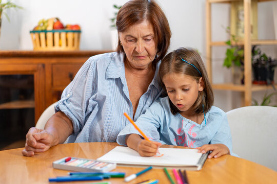 Grandmother watching girl drawing with colored pencil in book at home, showing encouragement and admiration while child focuses on creative activity  - Powered by Adobe