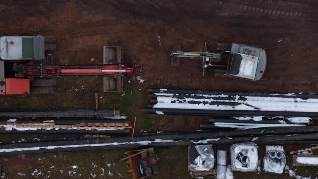 Top-down view of an industrial construction site showing excavators, stacked insulated pipes, and materials partially covered in snow. Earth-moving machinery is positioned near trenching.