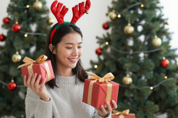 Joyful young Asian woman in festive reindeer antlers headband happily holds two Christmas gifts, celebrating the holiday season