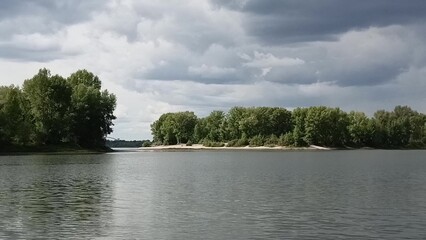 Ripples on the surface of a wide lake and a dense forest with tall bushes and trees on the opposite bank under a cloudy summer sky.