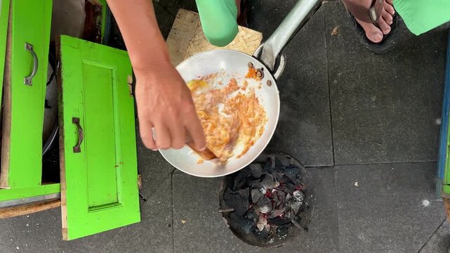 Top-down view of a street food vendor preparing Kerak Telor (traditional Jakarta omelette) with spiced rice and egg in a small wok over charcoal, showing hands and green stall.