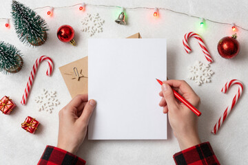 Top-down view of hands holding a blank Christmas card and red pen, surrounded by festive holiday decorations and lights, perfect for seasonal greetings and wish lists