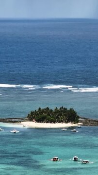 Island with palm trees and sandy beach surrounded by shallow coral reef and boats. Guyam Island. Siargao, Philippines.