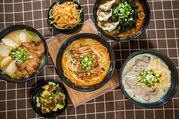 A top-down view of assorted Asian noodle soups and side dishes with dumplings, meats, vegetables, and various broths.
