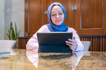 Indonesian muslim woman wearing a blue scarf is sitting at a table with a tablet in front of her. A concept of working in a cafe