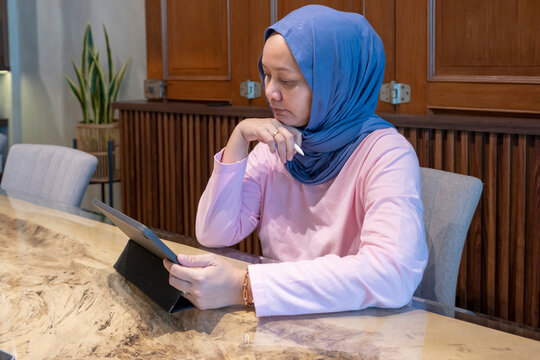 Indonesian muslim woman wearing a blue scarf is sitting at a table with a tablet in front of her. A concept of working in a cafe. She is focused on the tablet, possibly reading or working on it.