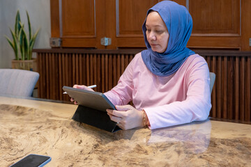 Indonesian muslim woman wearing a blue scarf is sitting at a table with a tablet in front of her. A concept of working in a cafe