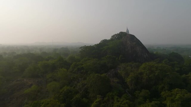 Side view of white dome temple crowning a rugged rock with jungle below in Sri Lanka.