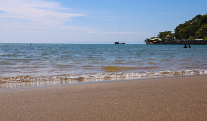 Coastal beach view with golden sand, ocean waves, and distant boat under bright blue sky