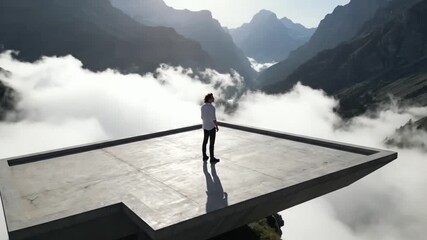 A man stands on a concrete platform, gazing at a breathtaking mountain landscape surrounded by clouds and valleys, a moment of awe