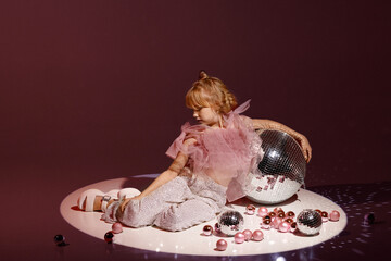 Studio shot of young Caucasian girl blonde, in festive outfit with sequin pants and pink top, sitting with disco balls and Christmas ornaments on burgundy background. Concept holiday, party.