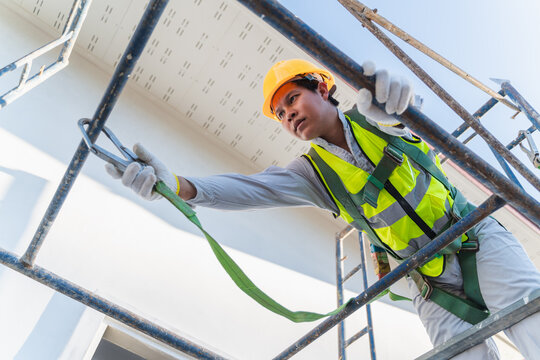 Construction worker in yellow helmet and safety gear climbing scaffolding, showcasing teamwork and safety in building site with clear blue sky