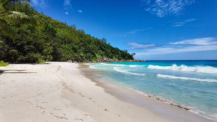 Tropical beach with footprints in the soft white sand, turquoise ocean waves, and lush green trees. Anse Georgette. Seychelles, Praslin.