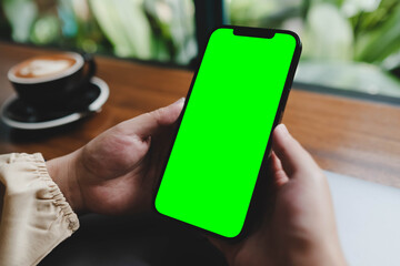 A person holds a smartphone with a bright green screen, perfect for chroma key editing, in a cafe setting with natural light.