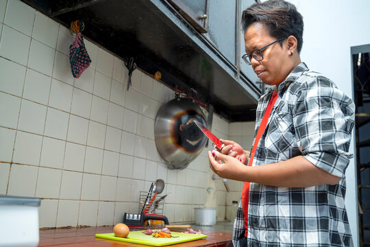 Indonesian southeast asian man standing in a kitchen peeling a red onion with a knife. Preparing ingredients on a cutting board