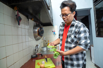 Indonesian southeast asian man standing in a kitchen preparing a meal. Holding green vegetables in the hand with chopped ingredients on a chopping board