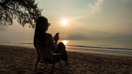 Woman relaxing and using her phone on a beach chair during a peaceful sunrise by the sea.
