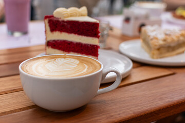 Taza de café latte con arte en espuma enfocada en primer plano, con rebanadas de pastel Red Velvet y tarta de manzana desenfocadas al fondo en una mesa de terraza.