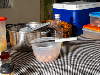 Outdoor food prep setup with pot, knife, vegetables, and containers on checkered tablecloth.