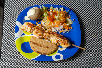 Grilled chicken skewer with salad, flatbread, and potato on colorful plate over checkered surface.