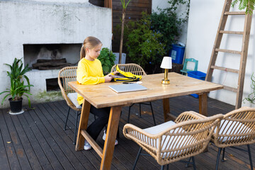 Young girl sitting at outdoor wooden table with backpack and laptop