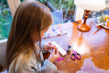 Young girl making beaded jewelry at a wooden table near a window