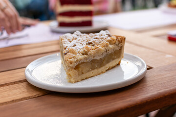 Primer plano de tarta de manzana con cubierta de crumble y azúcar impalpable. Al fondo se ve una torta Red Velvet y manos de personas, capturando un momento social en una cafetería al aire libre.