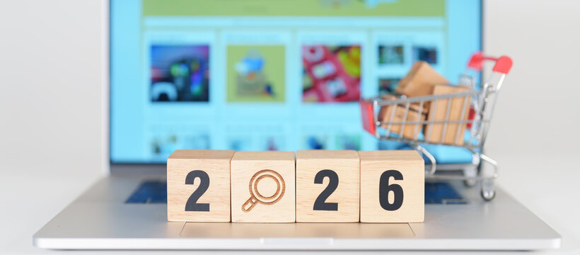 Happy New Year 2026 block with magnifying glasss and shopping trolley cart on table. E commerce, online shopping, finance, consumer economy and celebration concepts