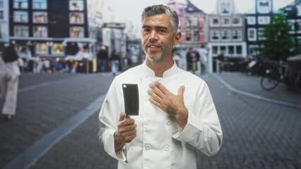 Man chef in white coat holds a cleaver in his right hand with left hand pressed to chest while standing on a cobbled street lined with buildings; pride gratitude.