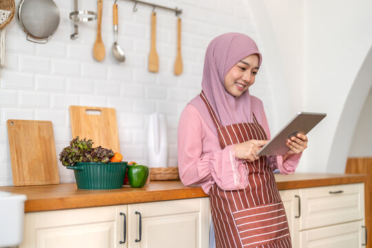 Smiling muslim woman wearing hijab use digital tablet in kitchen while preparing healthy food, online recipe search, home cooking and technology, halal cooking content, food blogger, healthy lifestyle