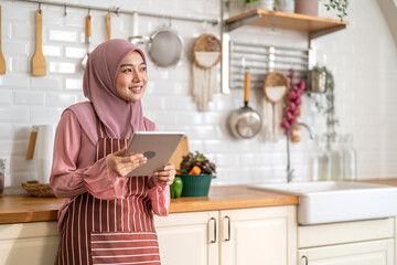 Smiling muslim woman wearing hijab use digital tablet in kitchen while preparing healthy food, online recipe search, home cooking and technology, halal cooking content, food blogger, healthy lifestyle