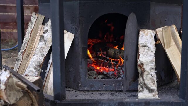 Outdoor wood stove burning birch logs, glowing embers inside black metal firebox, stacked split birch leaning at sides, thin smoke curling into cold air, rustic garden shed backdrop, close-up on flame