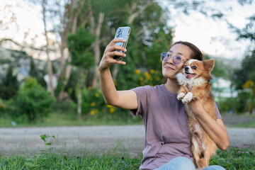 Happy Asian woman taking selfie with cute Chihuahua dog using smartphone in green public park.
