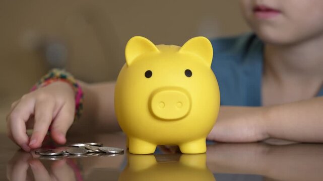 child placing coin into yellow pig bank on wooden table, focused hands, scattered coins, soft warm light,