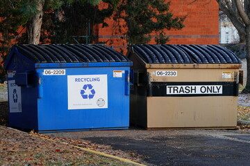 Two large commercial dumpsters, one blue for Recycling and one tan for Trash Only, sit side-by-side outdoors on a paved area with autumn foliage, symbolizing waste management.