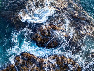 Aerial ocean texture. Turquoise waves from above. Crashing surf on rocks. Drone view of blue sea. Summer coastline aerial. Ocean foam top view. Rocky shore with waves. Tropical water texture.